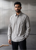 A man wearing a light gray long-sleeve button-down shirt and dark trousers stands in a minimalist studio with concrete walls and a window on the left.